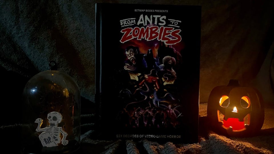 A picture showcasing the front cover of From Ants to Zombies, with a glowing ceramic pumpkin and skeleton diorama sitting on each side, against a soft grey backdrop.